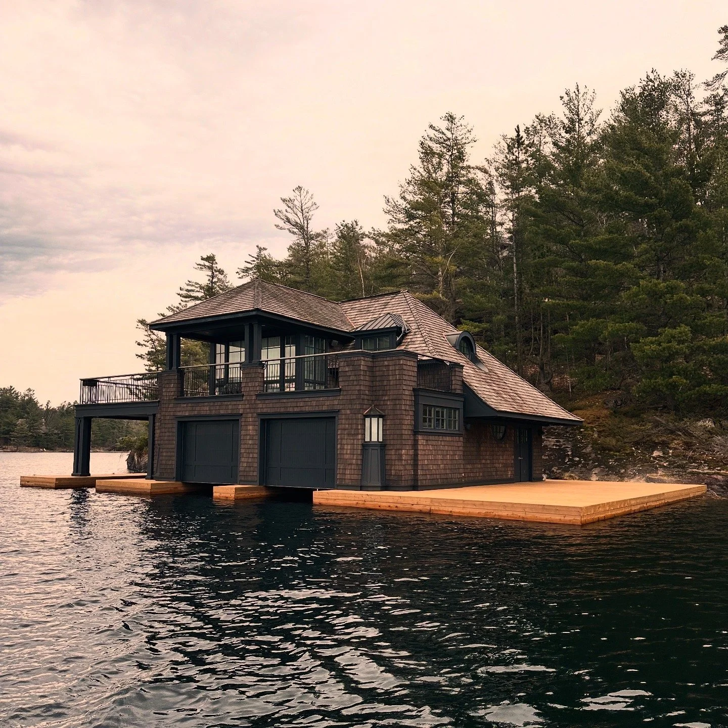 Cedar-shingled Muskoka boathouse with an upper terrace and timber docks, surrounded by lake water and pine forest at sunset